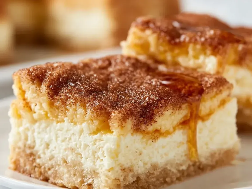 A square bar with a pale crescent-style dough base, a thick pale cheesecake centre and a cinnamon-sugar crusted top glistening with a honey drizzle