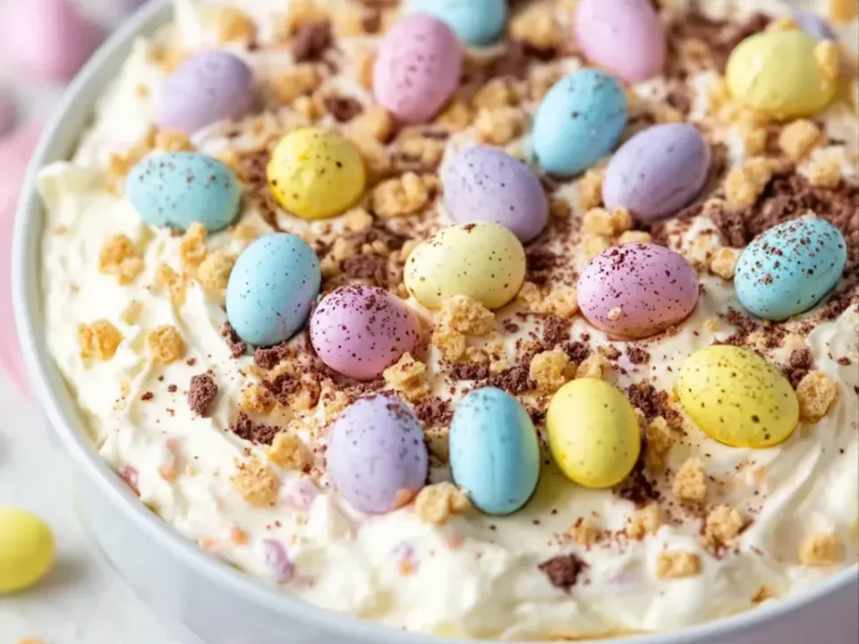 A thick soft ivory cheesecake dip in a white bowl topped with pastel speckled candy eggs, crushed golden biscuit crumbs and grated chocolate