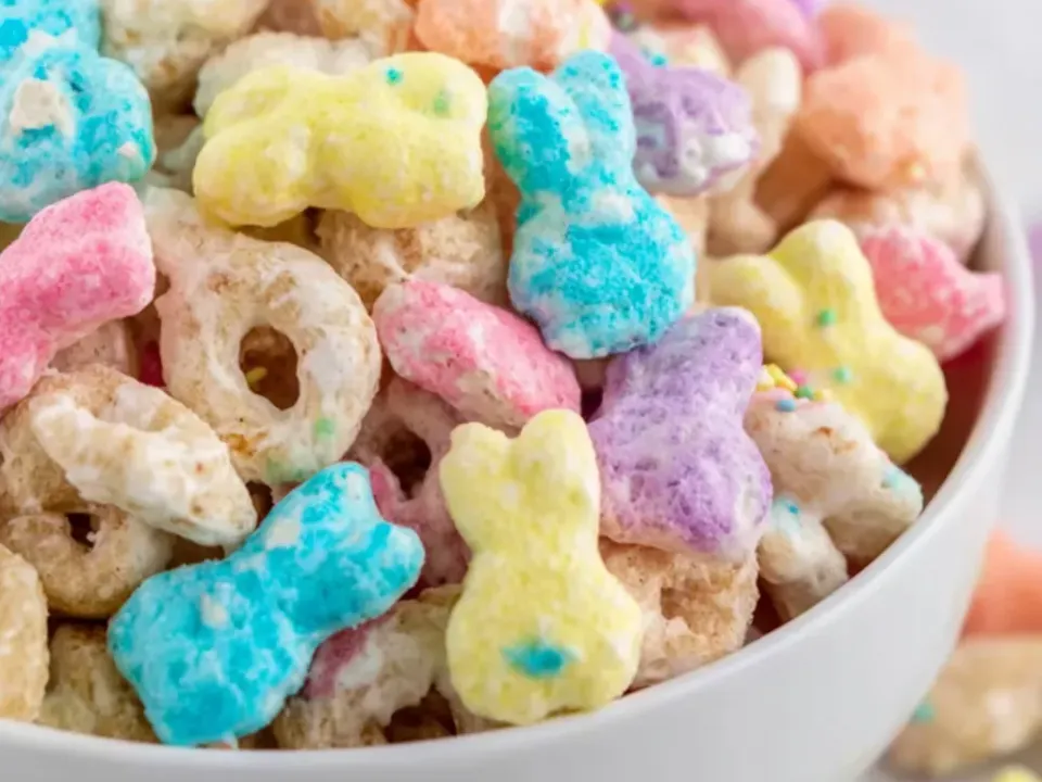 A white bowl heaped with toasted oat cereal rings and puffed cereal in a white coating, mixed with pastel bunny-shaped marshmallow candies and pastel sprinkles