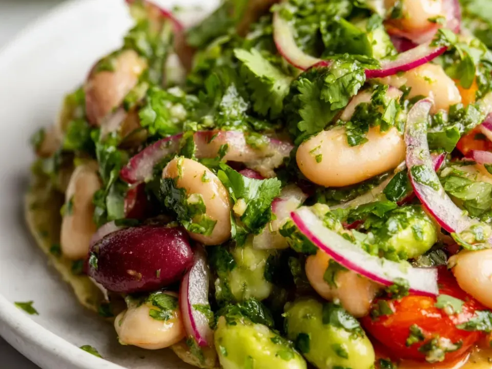 A cold salad of large white beans and dark red kidney beans with sliced red onion, cubed avocado and halved tomato, buried under heavily chopped cilantro in a green dressing with a lime wedge alongside