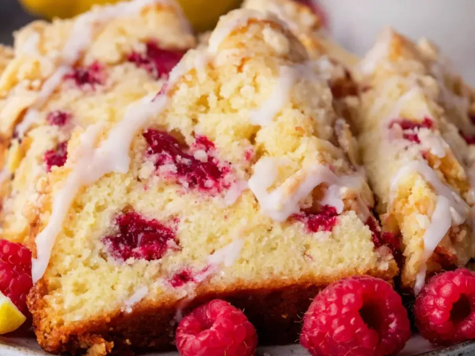 Thick slices of a loaf-shaped bake with a pale yellow crumb holding whole red raspberries, the top covered in coarse golden streusel and drizzled with thin white icing