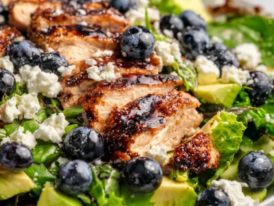 Sliced chicken with a dark caramelised balsamic-glazed surface over mixed greens in a white bowl, with whole blueberries, crumbled feta and diced avocado through the salad
