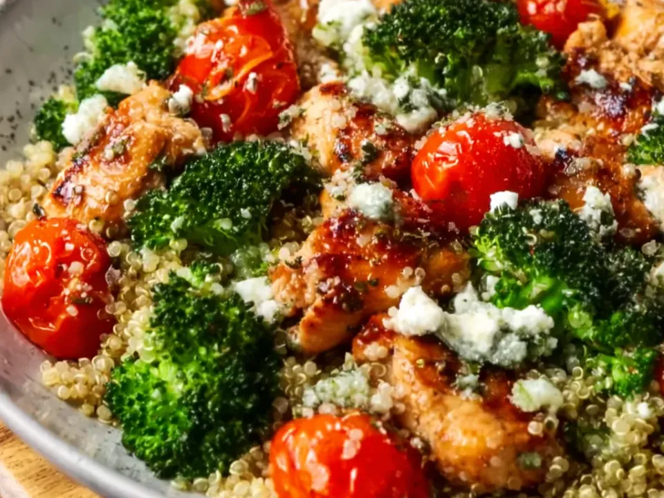 Chunks of seared chicken with browned edges over pale quinoa with bright green broccoli florets, whole blistered red cherry tomatoes and crumbled white feta, scattered with chopped parsley