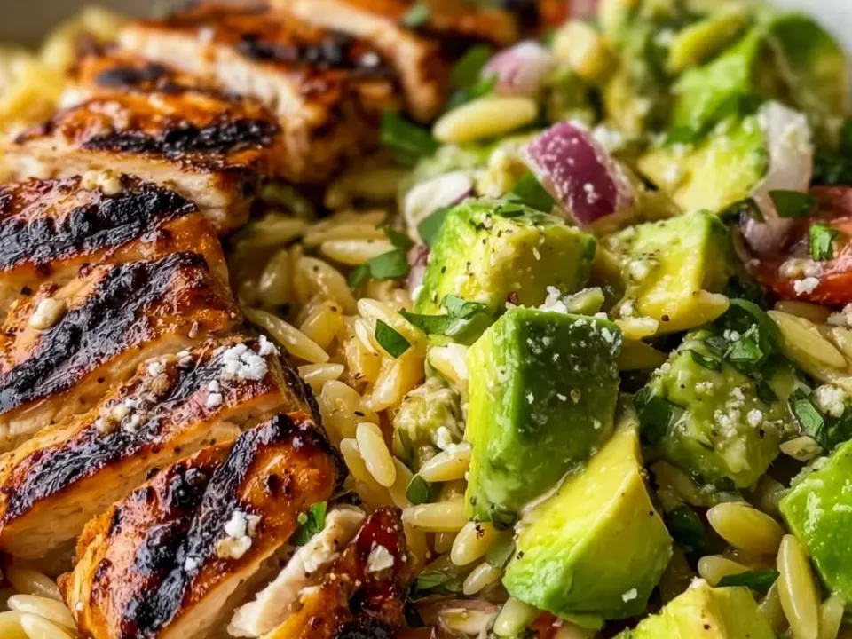 A bowl of orzo salad with sliced grill-marked chicken breast, crumbled feta, cubed avocado, red onion, halved cherry tomatoes and chopped herbs