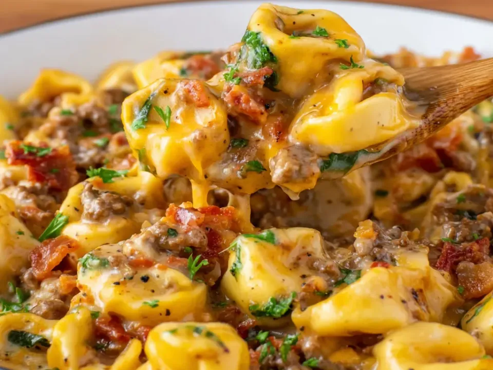A white bowl of cheese tortellini in an orange cheddar cream sauce with ground beef crumbles, crisp bacon pieces and parsley, one tortellini lifted on a wooden spoon with a cheese pull