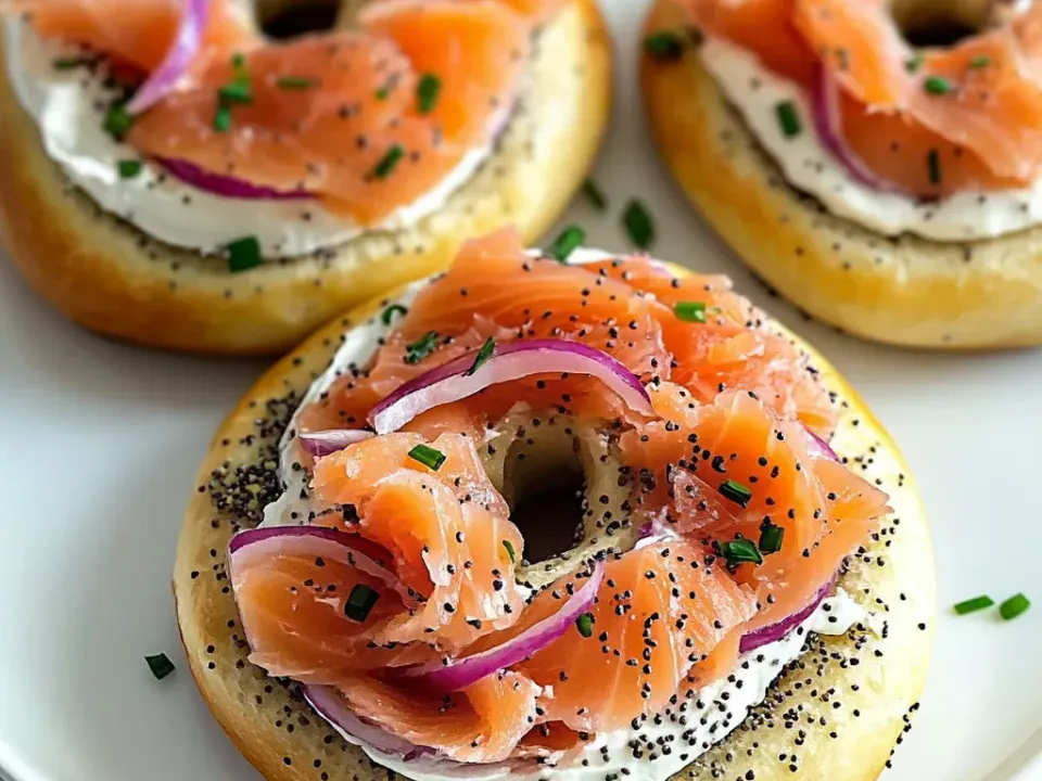 Halved poppy-seeded bagels spread with white cream cheese and topped with folded pale-pink cold-smoked salmon, raw red onion rings and chopped chives on a white plate