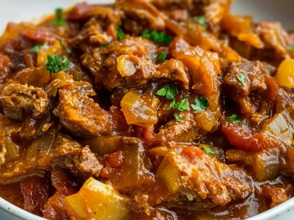 A white bowl of thick braised stew with chunks of browned meat, soft onion and cabbage strips and tomato in a glossy red-brown paprika gravy, garnished with parsley
