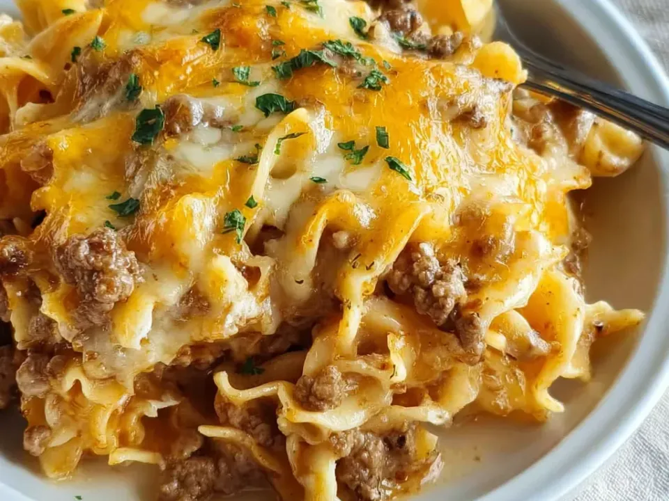 A white bowl of wide egg noodles and browned ground beef in a light creamy sauce under melted orange cheddar and white cheese, sprinkled with parsley