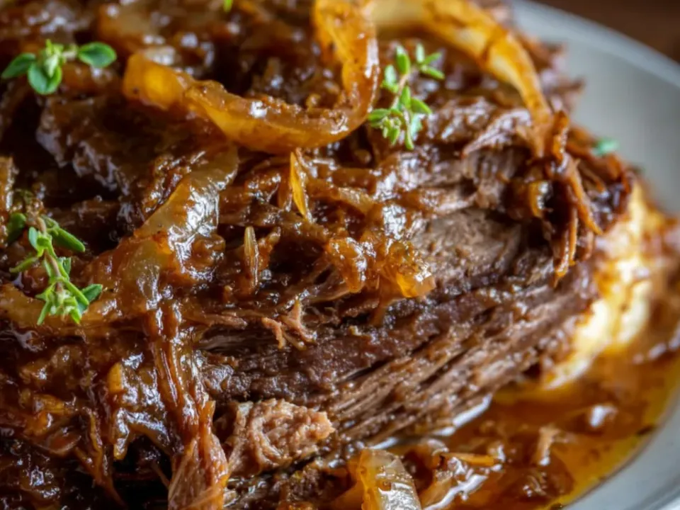 Pull-apart beef roast on a grey plate under dark caramelised onions and thick brown gravy with thyme, mashed potato beneath