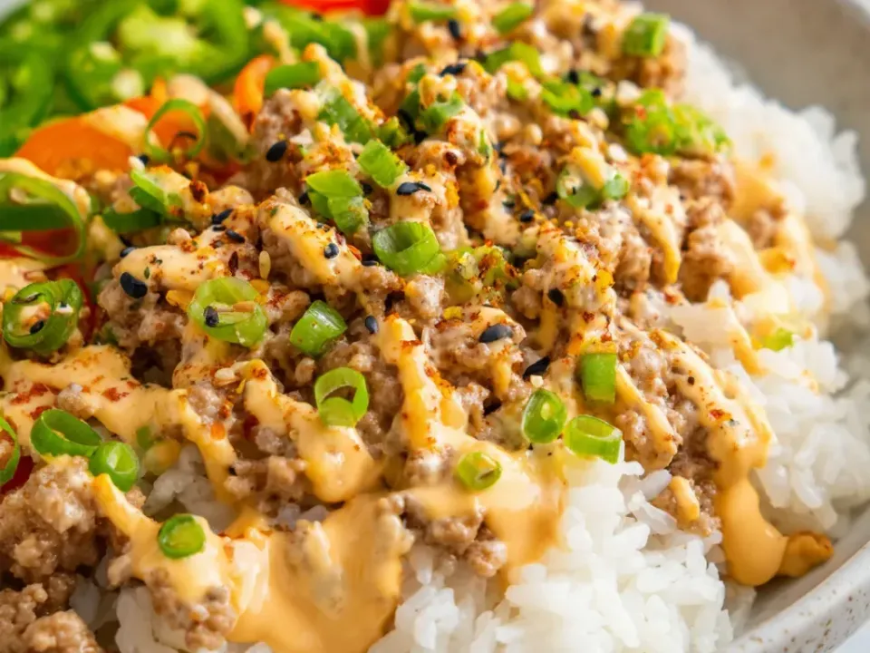 A bowl of white rice topped with browned ground turkey crumbles, a thick creamy orange-pink sauce, sliced green onion, black and white sesame seeds and sliced green and red chilies