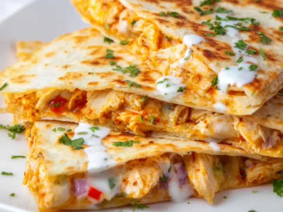 Stacked buffalo chicken quesadilla wedges on a white plate with griddled blistered tortillas, melted cheese, diced red onion and pepper and a white ranch drizzle