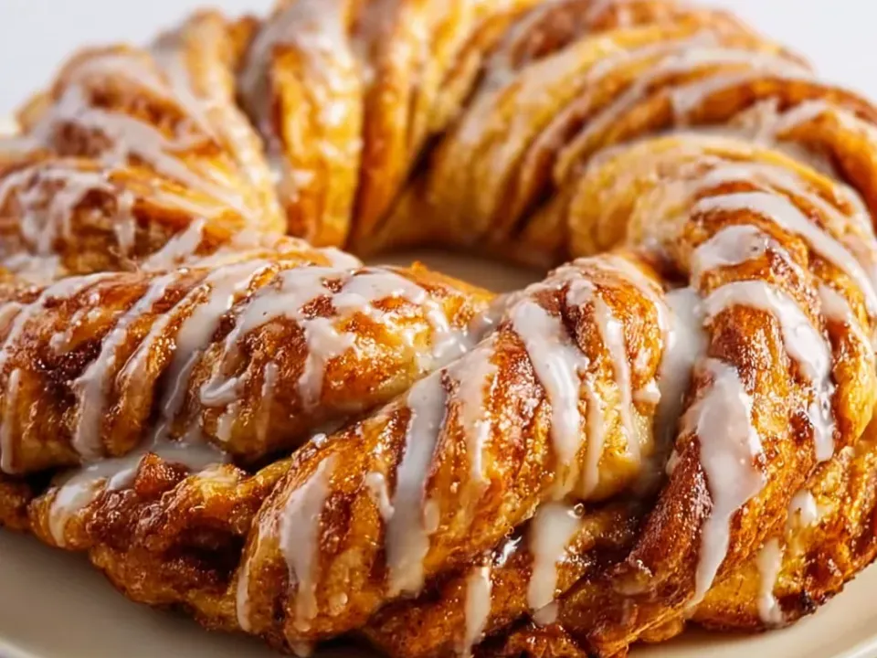 A large twisted ring of golden laminated pastry with visible layered folds, drizzled all over with a thin white glaze, on a cream plate