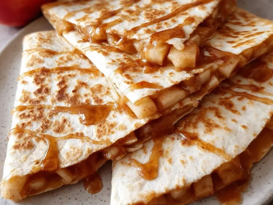Wedges of flour tortilla quesadilla toasted with brown spots and dusted with cinnamon, filled with diced cooked apple in syrup and drizzled with caramel on a speckled plate
