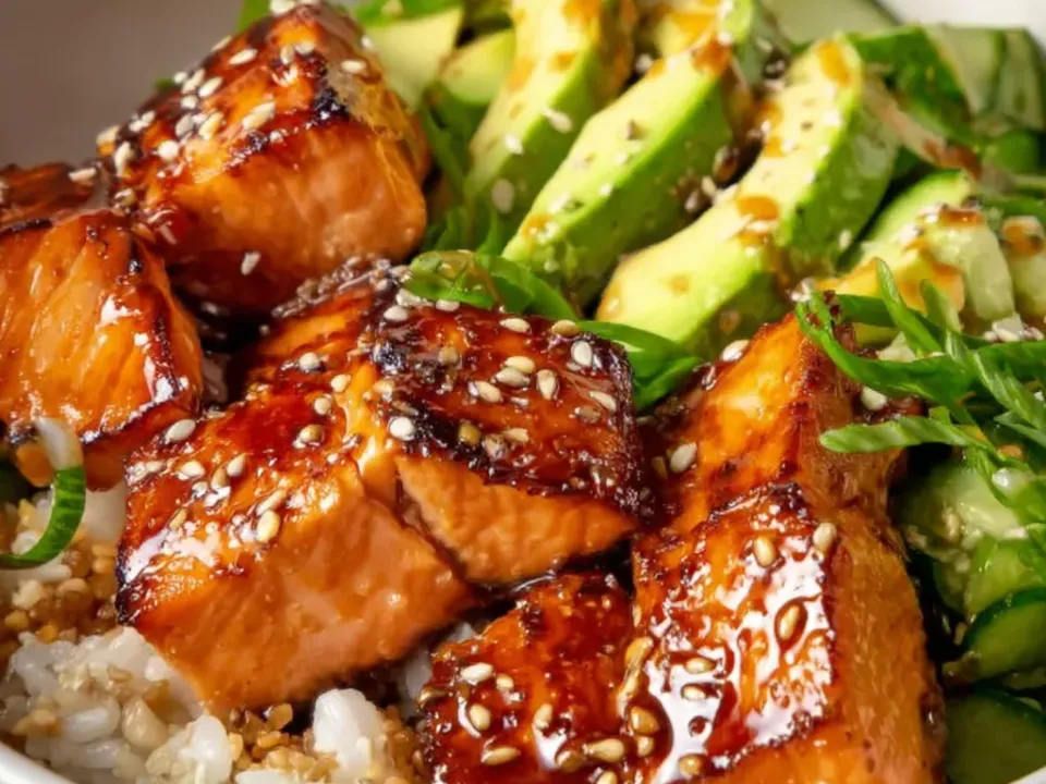 Cubes of salmon with deeply caramelised dark amber glazed surfaces scattered with sesame seeds, over white rice with sliced avocado, sliced cucumber and shredded green leaves