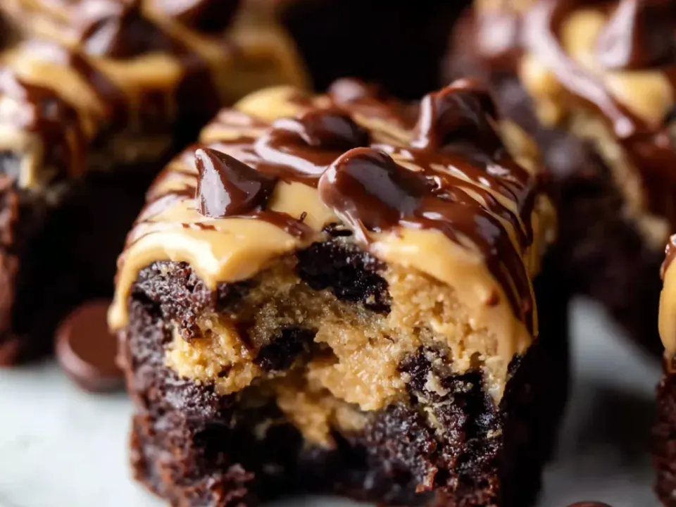 Individual bite-sized brownies topped with a dollop of cookie dough, one bitten into showing the fudgy base