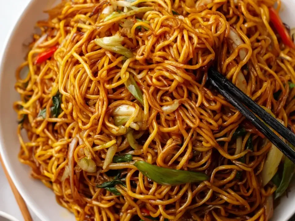 A white platter of thin glossy brown stir-fried egg noodles with green onion lengths, shredded cabbage, red pepper strips and greens, lifted with black chopsticks