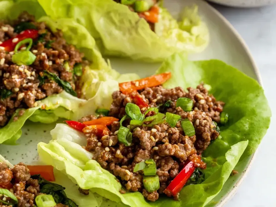 Cupped butter-lettuce leaves on a plate filled with browned seasoned ground beef, strips of red pepper, sliced green onion and sesame seeds