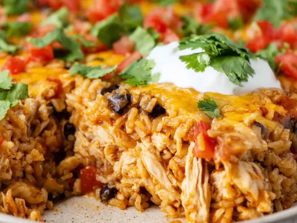 A cut portion of Mexican chicken and rice casserole with black beans and melted cheese, topped with diced tomato, cilantro and soured cream