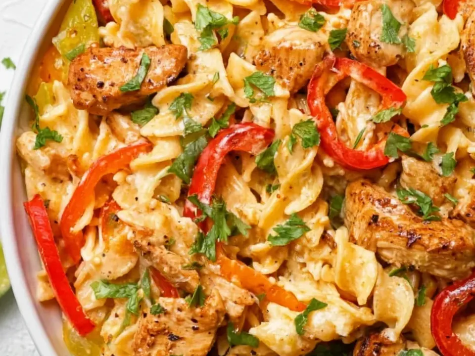 Bowtie pasta in a creamy pale orange sauce with seared seasoned chicken and long red and orange bell pepper strips, heavy with chopped cilantro and lime wedges