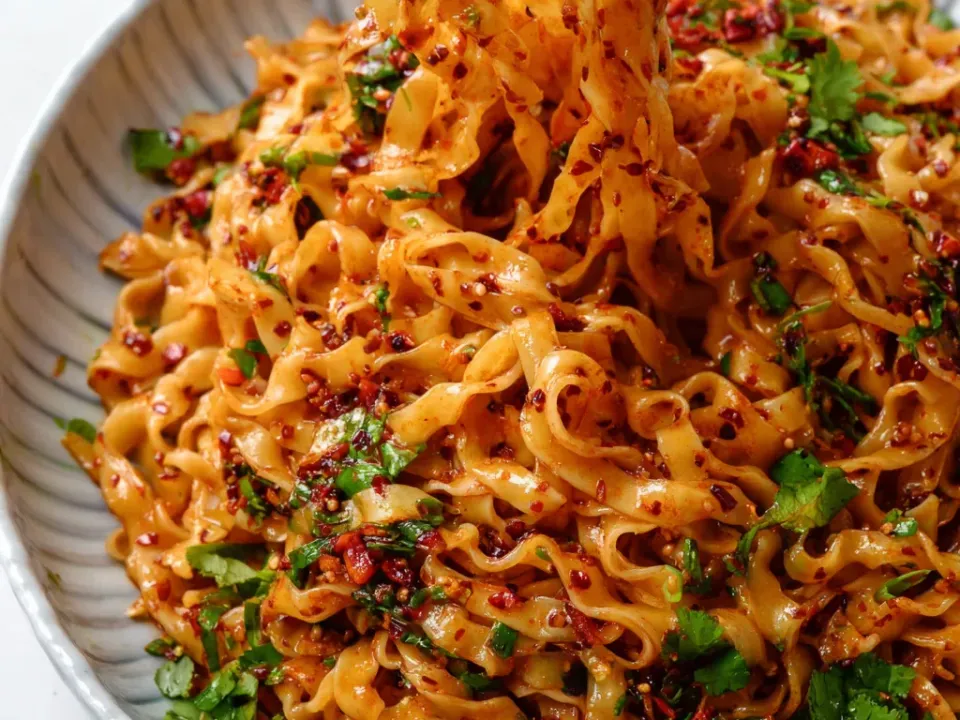 Wide wheat noodles glossy with chilli oil, flecked with red chilli flakes, sesame and chopped coriander, lifted on chopsticks