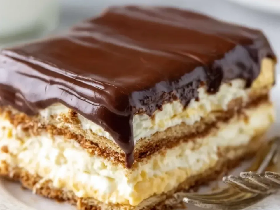 A square of no-bake eclair cake on a white plate showing graham cracker layers alternating with thick pale vanilla cream under a glossy dark chocolate ganache top