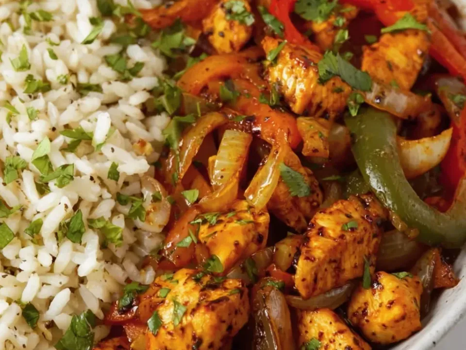 A bowl of spiced seared chicken chunks with red and green bell pepper strips and softened onion, garnished with cilantro, beside white rice mixed with chopped cilantro