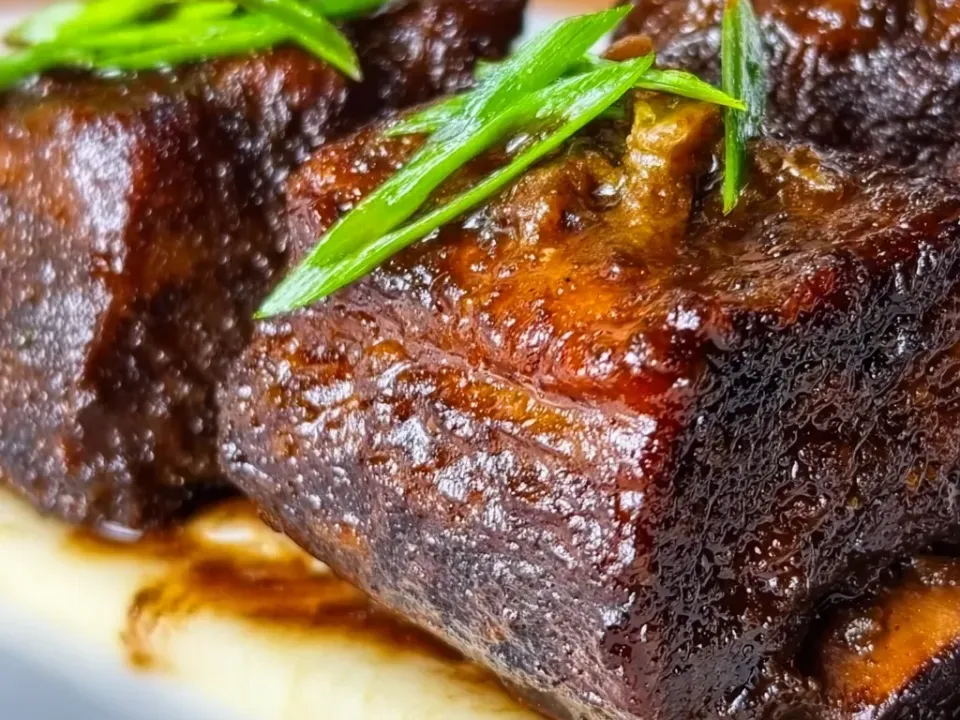 Thick pieces of dark glazed braised beef short rib topped with long slices of green onion, resting on a bed of pale mashed potato