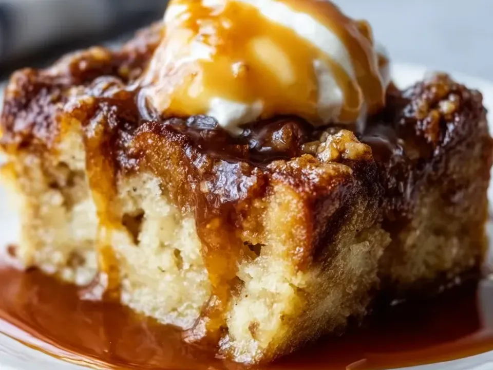 A tall square of bread pudding with a dark caramelized top and pale custard crumb in a pool of amber toffee sauce, topped with a scoop of vanilla ice cream under more sauce