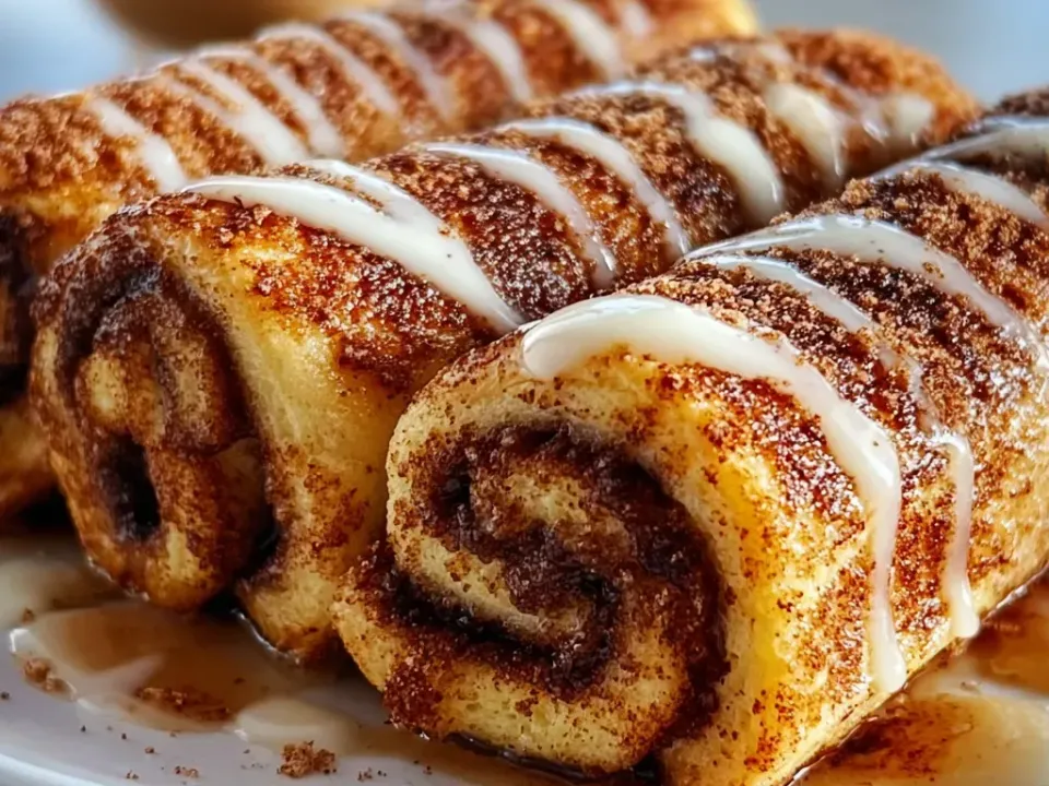 Log-shaped roll-ups of flattened white bread griddled golden and coated in dark cinnamon sugar, cut ends showing a tight brown spiral, drizzled with white icing over a pool of amber syrup